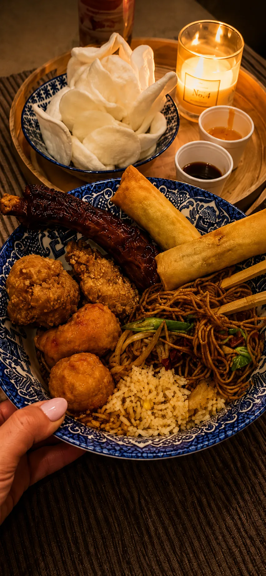 A hand holding a willow-pattern plate of chow mein, salt & pepper balls and a spring roll with prawn crackers